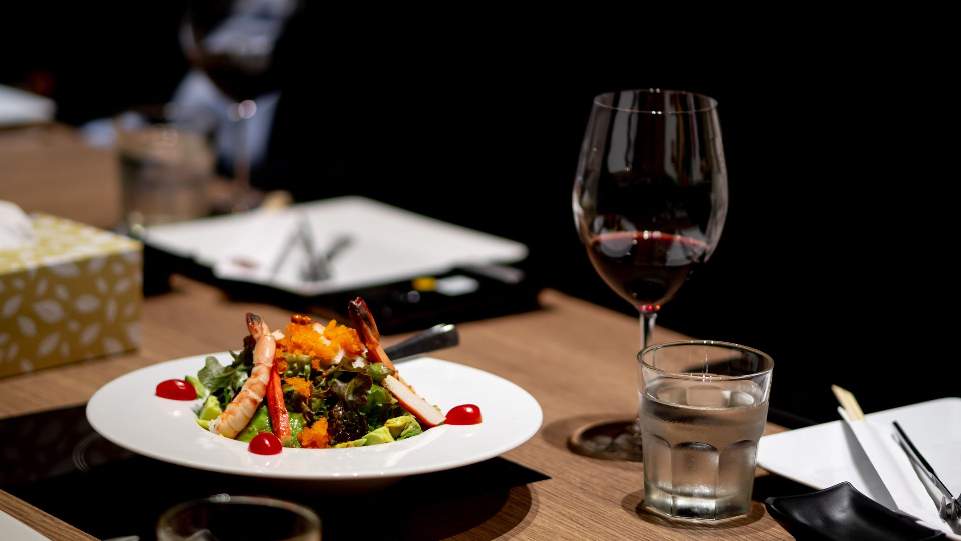 A vibrant salad with shrimp and colorful toppings graces the plate, accompanied by a glass of red wine and water on a wooden table. Discover this delightful setting at one of the charming restaurants in Playa del Coco, where culinary artistry meets tropical ambiance.